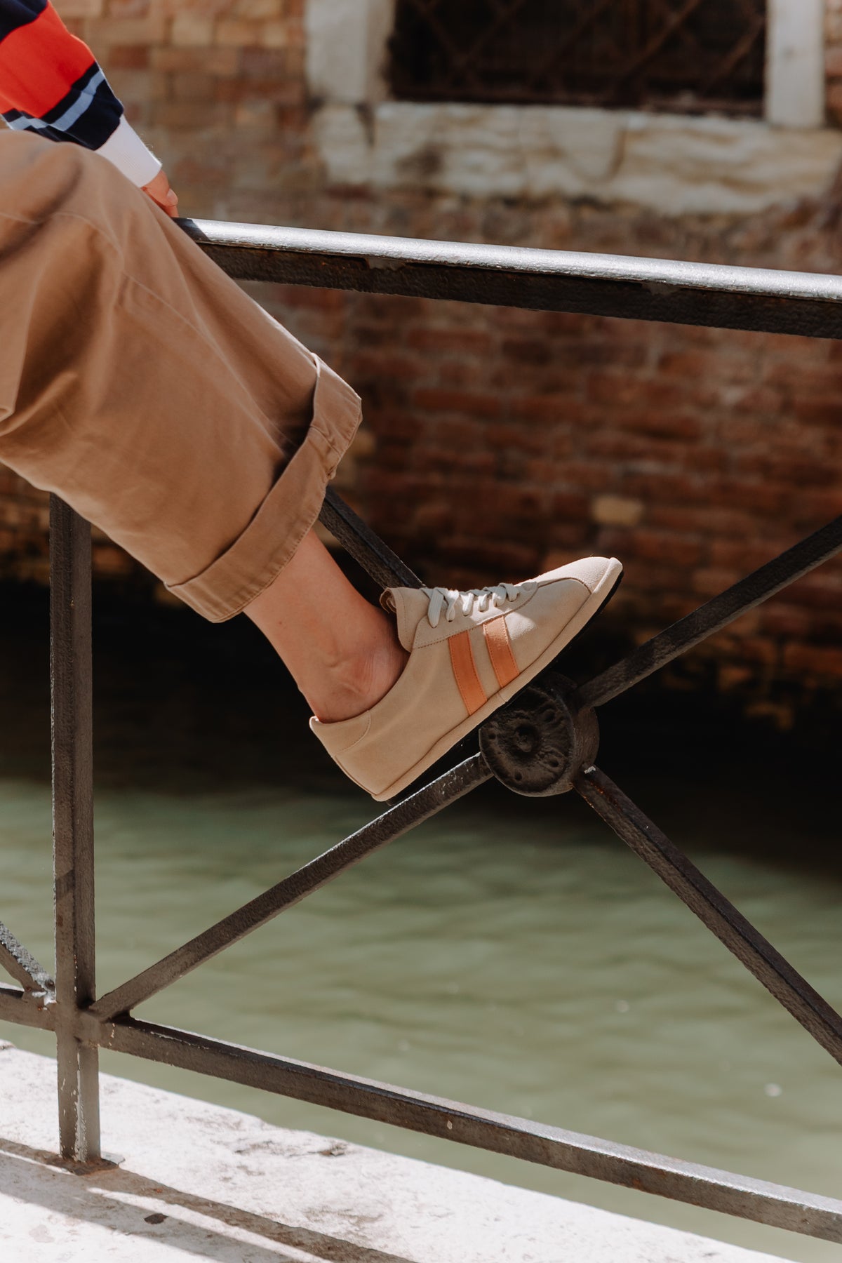 A beige suede gondolina sneaker with two vertical peach-orange stripes on the side, matching beige laces, worn by model, detail